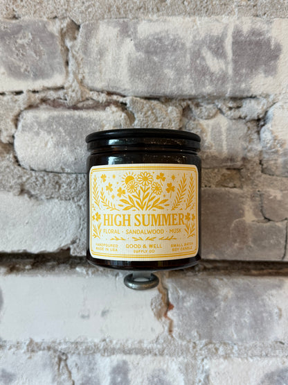 Folk & Flame High Summer Soy Candle in a clear glass jar with a wooden wick, surrounded by summer-themed decor.