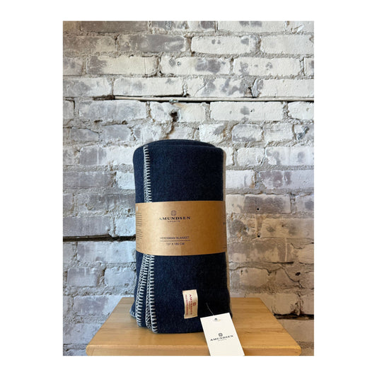 Amundsen Herdsman Blanket in faded navy, cozy wool accessory with fringed edges, draped on a rustic wooden surface.
