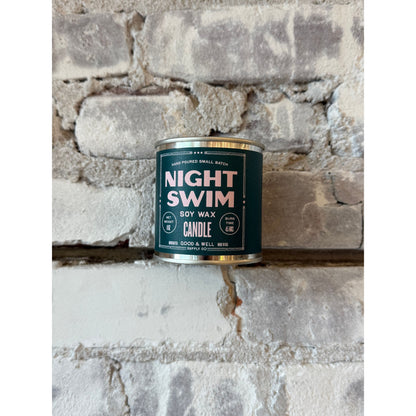 Night Swim soy candle by Good & Well Supply Co. in a glass jar, with a lit wick and a calming, dark blue label design.